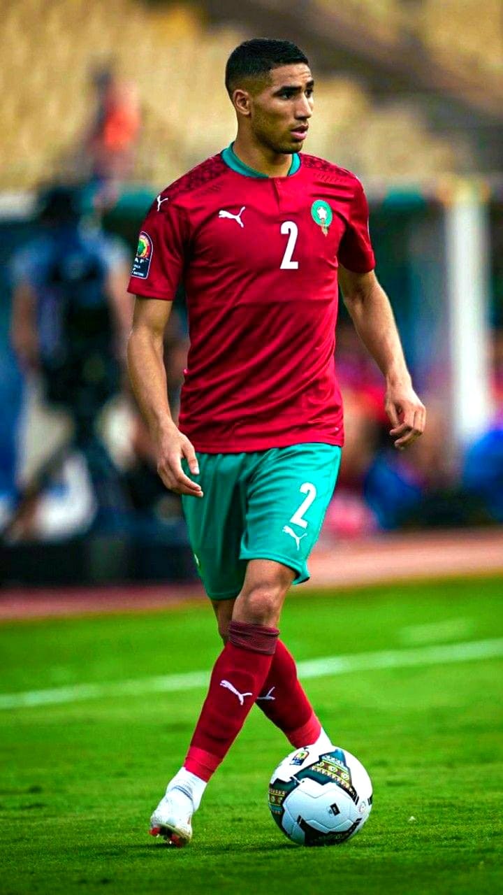 A soccer player in a red Morocco jersey with the number 2, dribbling a ball on a grass field during a match, as seen on IPTV Suisse.